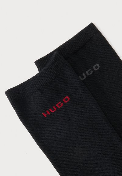 Paire de chaussettes noires avec bords-côtes, présentant le logo "HUGO" brodé près du haut en fil rouge et gris foncé sur chaque chaussette.