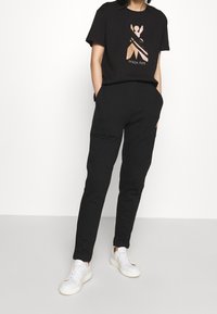 Camiseta de algodón negra con diseño gráfico metálico; pantalones de chándal negros con bolsillos laterales; zapatillas blancas. Atuendo casual con un corte relajado.