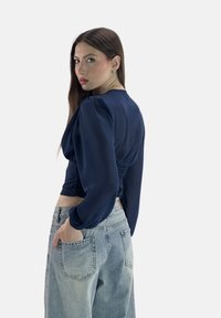Navyblaues, gekürztes Oberteil mit puffigen langen Ärmeln und gerafftem Rücken, kombiniert mit hellblauen, hochgeschnittenen Jeans. Glatte Stoffe, minimale Details.