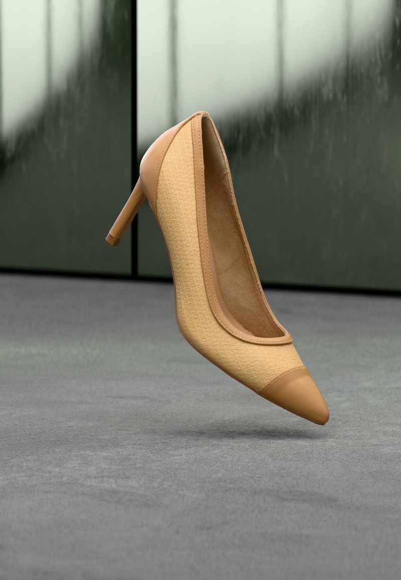 Chaussure à talons hauts beige avec bout pointu et tissu texturé, flottant au-dessus d'une surface grise avec un arrière-plan vert flou.