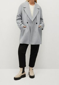 Szary, dwurzędowy płaszcz z mieszanki wełny z dużymi klapami, dwoma czarnymi guzikami, kieszeniami po bokach, noszony na kremowym swetrze i czarnych spodniach.