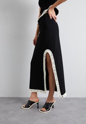 Kobieta w czarnej midi spódnicy z białą ozdobną lamówką i wysokim rozcięciem na udzie, zestawiona z czarnymi sandałami na wysokim obcasie wsuwanymi.