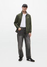 Olajzöld cipzáras dzseki, fehér póló, megkopott fekete farmernadrág és fekete sportcipő. A dzsekin magas gallér és diszkrét logó részlet található.