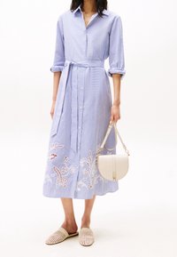 Donna che indossa un abito midi a righe blu e bianche con ritagli floreali, mentre tiene una borsa beige ed è vestita con scarpe slip-on intrecciate beige.