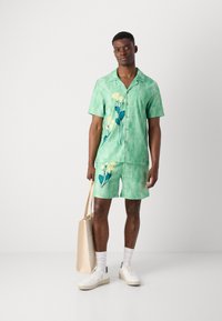 Grøn kortærmet skjorte og matchende shorts har et tie-dye mønster og gul blomsterbroderi. Modellen holder en beige tote bag.