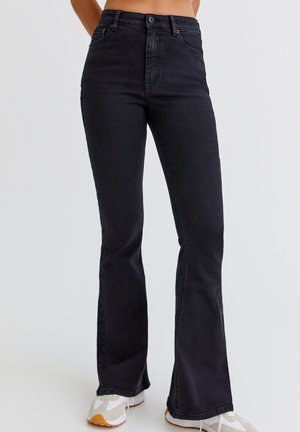 Schwarze, hochtaillierte Schlagjeans, getragen mit weißen und beigen Sneakern, von der Taille bis zu den Füßen an einem neutralen Hintergrund präsentiert.