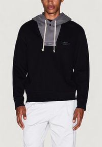 Joven vestido con un sudadera negra con capucha gris y cuerdas de ajuste, combinada con pantalones de color claro, frente a un fondo liso.