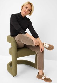Grüner Bezugssessel mit geschwungenem Design; Person in einem schwarzen Rollkragenpullover, beigen Cordhosen und braunen Sneakers mit weißen Schnürsenkeln.