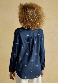 Woman with curly hair wearing a navy blue long-sleeve blouse with colorful car prints, standing against a plain beige background.
