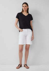 Femme portant un haut noir à manches courtes en V, un short blanc à ourlet roulé de longueur genou et des sandales noires, se tenant contre un mur gris uni.