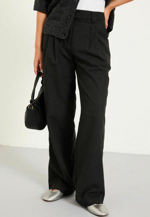 Weite schwarze Hose aus strukturiertem Stoff, mit Falten, einem Bund mit Knöpfen und kombiniert mit silbernen Ballerinas und einer schwarzen Handtasche.
