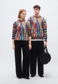 Pulls en tricot avec des motifs abstraits multicolores, associés à un pantalon noir à jambes larges. Un sac à main noir circulaire avec une petite poignée.