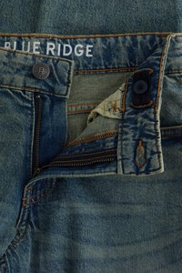 Detail modrých riflí s čiastočne otvoreným zipsom, gombíkom a značkovou etikietou "BLUE RIDGE" na vnútornej časti opaska.