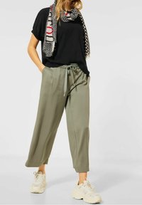 Top nero a maniche corte abbinato a pantaloni larghi verde oliva, con vita elastica e una sciarpa decorata. Calzature leggere completano l'outfit.
