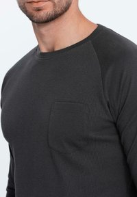 Uomo che indossa una maglietta grigia scura a maniche lunghe con una piccola tasca sul petto, che mostra i dettagli della parte inferiore del viso e del collo.