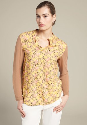 Bluse mit floralem Muster und gelbem Hintergrund, verziert mit rosa Rosen und grünen Akzenten. Lange braune Ärmel, Kragen und eine lockere Passform.