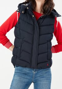 Kvinna i en marinblå qiltad väst med framfickor, över en röd långärmad topp och blå jeans, med händerna vilande på höfterna.