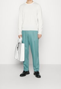 Persona vestida con una camiseta de manga larga blanca, pantalones turquesa, zapatos negros y sosteniendo un gran bolso blanco contra un fondo liso.