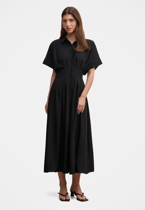 Femme aux longs cheveux bruns portant une robe midi noire boutonnée à manches courtes et des sandales noires à talons, debout devant un fond uni.