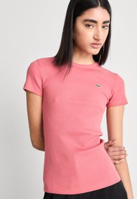 Rožinė gipiūrinė t-shirt marškinėliai su trumpomis rankovėmis, apvaliu kaklu ir mažu žaliu logotipu kairėje krūtinės pusėje. Minkštas medvilnės audinys, aptemptas dizainas.