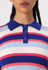 Polo rayé avec un col bleu. Présente une texture côtelée, quatre boutons bleus et des rayures alternées en bleu, crème et rouge.