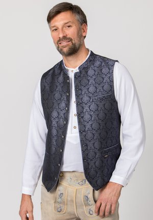 Mann trägt ein weißes Hemd, eine dunkelblaue Brokatweste und beige traditionelle bestickte Lederhosen vor schlichtem Hintergrund.