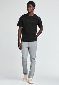 Svart bomullst-shirt med texten "Polo Club", kombinerad med ljusgrå byxor och beige sneakers, stående mot en ljus bakgrund.