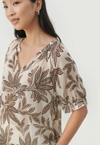 Beige bluse med brune blomsterprint, V-udskæring og korte puffærmer. Fremstillet af letvægts, semi-gennemsigtigt stof med subtil drapering.