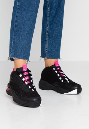 Person iført sorte sneakers med pink og hvide detaljer og blå jeans med flossede kanter, der står på et hvidt gulv.