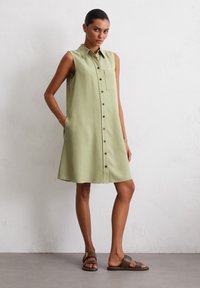 Robe sans manches en tissu vert clair avec un devant à boutons, un col pointu et une poche poitrine unique. Portée avec des sandales marron.