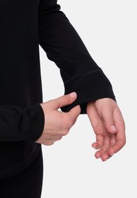 Schwarzes Langarm-Oberteil mit einer taillierten Passform. Nahaufnahme einer Hand, die den Bund anpasst, wobei der glatte Stoff und die kontrastierenden Stichdetails sichtbar sind.