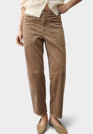 Vrouw draagt hoge taille bruine broek met voorzakken en beige platte schoenen, staand tegen een effen achtergrond.
