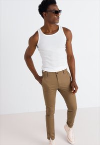 Pantalon marron léger avec une coupe slim, fermeture à bouton et poches latérales, associé à un débardeur blanc et des baskets blanches.