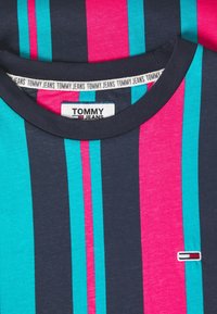 Detailní záběr na pruhovanou košili s námořnickými, tyrkysovými a růžovými vertikálními pruhy, s označením a logem Tommy Jeans blízko límce a na hrudi.