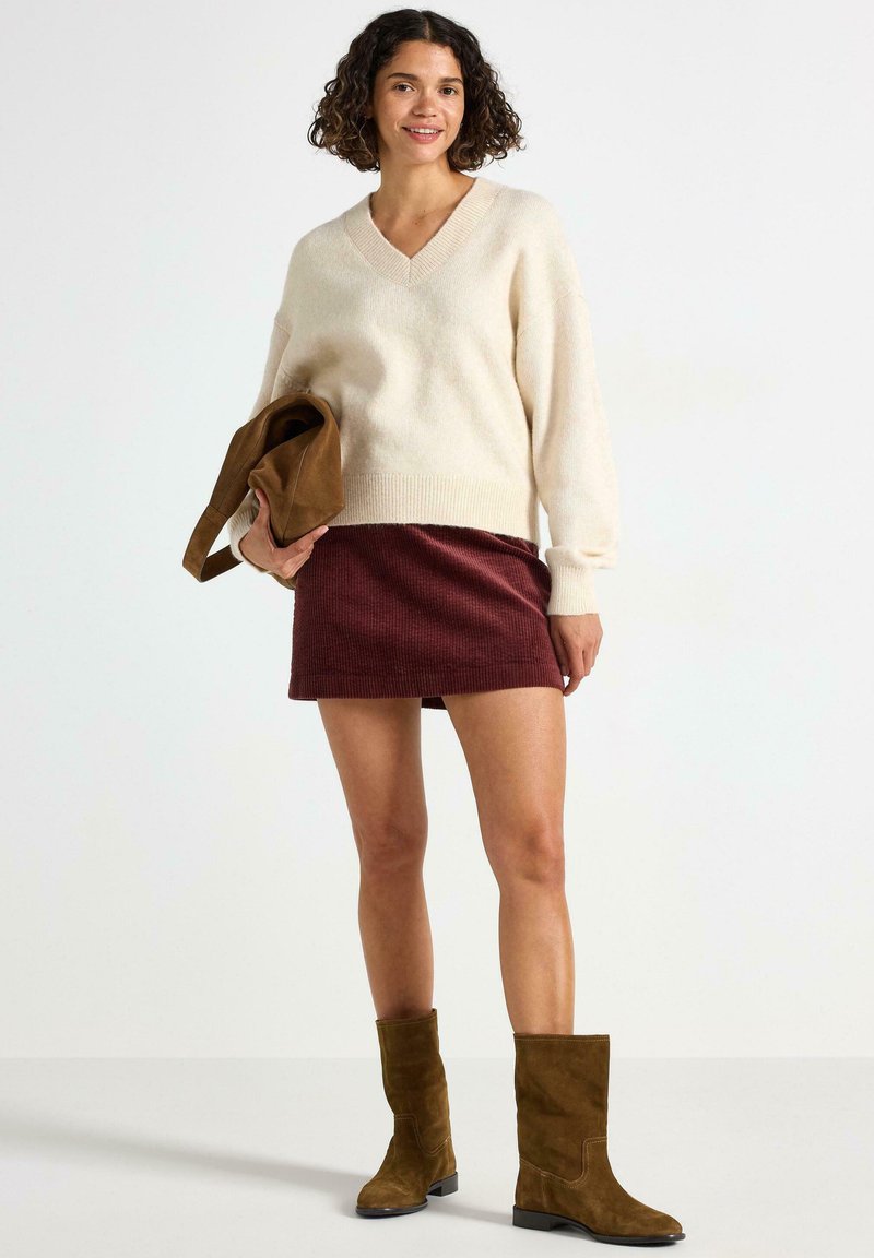 Beige v-hals strikket sweater, bordeaux ribbet mini nederdel og brune ruskinds ankelstøvler med blokhæle. Model holder en brun clutch.