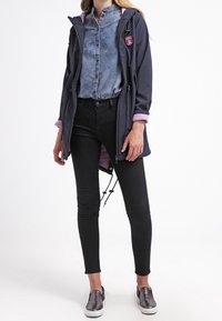 Vrouw in een marineblauwe jas met een roze voering, denim blouse, zwarte skinny jeans en metallic instappers, staande voor een witte achtergrond.