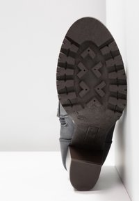 Semelle de botte noire avec un motif de crampons robustes et un talon bloc, positionnée à la verticale contre une surface blanche.