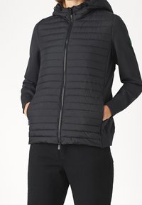 Persona que lleva una chaqueta negra acolchada con cierre y capucha, con las manos en los bolsillos, combinada con pantalones negros frente a un fondo blanco liso.