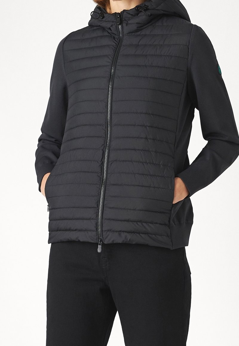 Persona que lleva una chaqueta negra acolchada con cierre y capucha, con las manos en los bolsillos, combinada con pantalones negros frente a un fondo blanco liso.