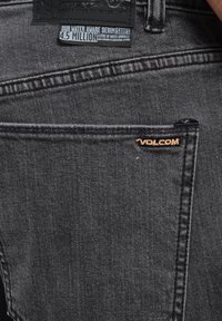 Donkergrijze denim jeans hebben een achterzak met een klein Völcom-logo in lichtbruin en een label dat de waterbesparende productiemethoden beschrijft.