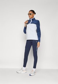 Fleece genser i hvit og marineblå, med en halv-glidelås krage. Marineblå leggings med cargo-lommer og blå glidelåser. Grå joggesko med detaljer.