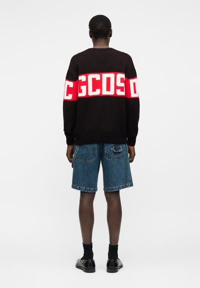 Άνδρας όρθιος, βλέποντας προς τα πίσω, φοράει μαύρο πουλόβερ με λευκή και κόκκινη ταινία λογότυπου "GCDS", μπλε τζιν σορτς, μαύρες κάλτσες και μαύρα παπούτσια.