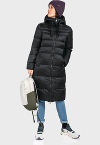 Schwarze Pufferjacke mit Kapuze, Reißverschluss und gestepptem Design. Kombiniert mit hellblauen Jeans, einer lila Mütze und einem weiß-grünen Rucksack.