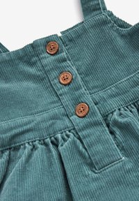 Miętowa sukienka z kordurowego materiału ma regulowane ramiączka, trzy brązowe guziki oraz marszczenia w pasie, które dodają faktury.