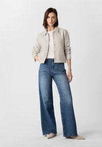 Hellgraue Button-up-Jacke mit zwei Seiten Taschen, darunter ein weißes Oberteil, kombiniert mit weit geschnittenen blauen Jeans und beigefarbenen spitzen Ballerinas.