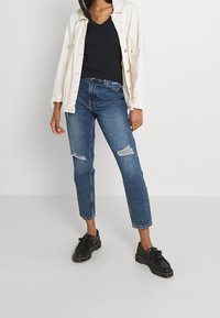 Ljus krämfärgad skjorta med knappar över en svart v-ringad topp, tillsammans med slitna blå jeans och svarta loafers med fransdetaljer.