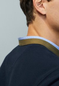 Läheltä kuva henkilön kaulasta ja korvasta, joka on pukeutunut tummansiniseen paitaan, jossa on vihreä ja vaaleansininen kaulus.