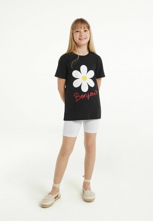 T-shirt en coton noir avec un grand graphique de marguerite blanche et le texte "Bonjour" en rouge. Associé à des shorts blancs et des espadrilles beiges.