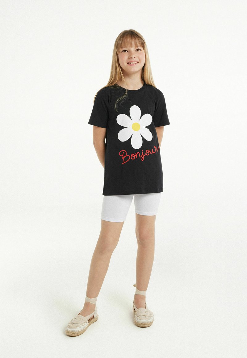 T-shirt di cotone nera con una grande grafica di margherita bianca e testo rosso "Bonjour". Abbinato a pantaloni corti bianchi e espadrillas beige.