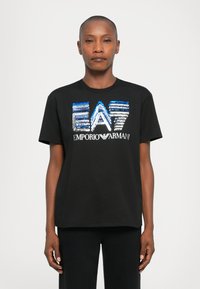 Cilvēks ar īsiem matiem, ģērbies melnā T-kreklā ar zilu un sudraba mirdzoša "EA7 Emporio Armani" logo uz vienkārša balta fona.